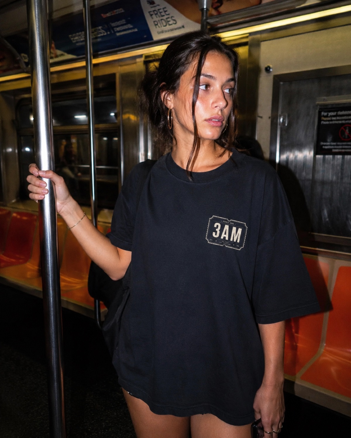 Woman in the 3AM boxed-logo tee holding the pole on a late-night subway
