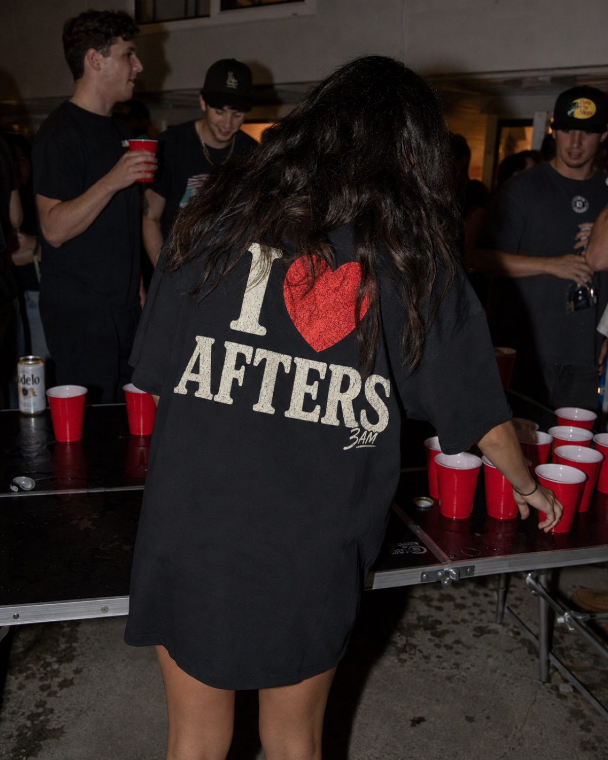 Woman from behind in the I heart Afters tee playing beer pong at a house party