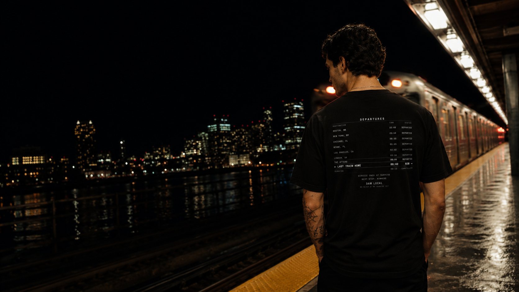 A figure in the Last Train Home departures tee on a Boston train platform at night, city skyline lit across the water